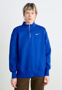 Sudadera Nike azul con cuello alto y media cremallera, fabricada en un material suave. Presenta mangas largas con puños acanalados y un pequeño logo.