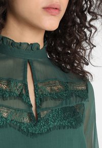 Grön genomskinlig blus med en vågig hög halsringning, spetsdetaljer och en liten nyckelhålsdetalj framtill. Diskret plissering och texturiserad tyg.