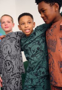 Trois enfants portant des T-shirts imprimés. Un gris avec des motifs noirs, un vert avec des crânes, et un marron avec des accents orange.