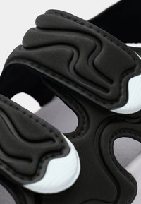 Μαύρο σανδάλι με υφασμένα λουριά διακοσμημένα με κυματιστά σχέδια και λευκές λεπτομέρειες. Ο σχεδιασμός περιλαμβάνει ανοιχτά τμήματα για αερισμό.