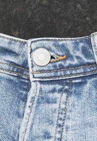 Jeans de ganga azul claro com fecho em botão prateado, costura laranja em contraste e padrões de textura visíveis.