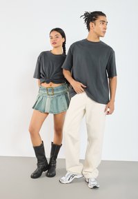 Two models stand side by side. The woman wears a dark gray cropped t-shirt, a pleated denim skirt, and black knee-high boots. The man wears a dark gray t-shirt, off-white loose trousers, and silver sneakers.