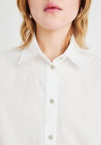 White button-up shirt with a classic collar, featuring five buttons, lightweight fabric, and a slightly wrinkled texture.