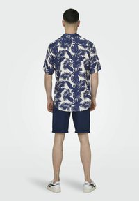 Camicia a maniche corte, con bottoni, caratterizzata da un motivo di foglie di palma navy su sfondo crema, abbinata a pantaloni corti navy arrotolati e sneakers bianche.