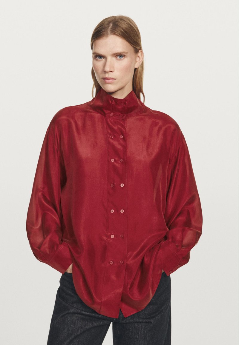 Red satin shirt with a high collar, button-down front, and loose fit. Long sleeves with relaxed cuffs; smooth, shiny texture.