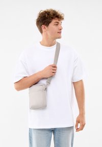 Hellgraue Umhängetasche mit rechteckiger Form, Reißverschluss und verstellbarem Tragegurt, getragen von einer Person in einem weißen T-Shirt.