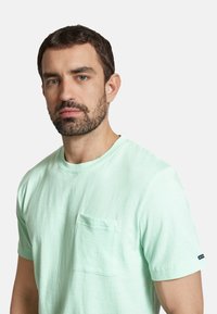 Tricou din bumbac de culoare verde mentă deschis, cu mâneci scurte, decolteu rotund și un buzunar frontal. Model texturat subtil pe material.