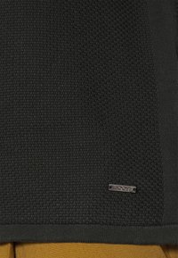 Tissu en maille noire texturée avec une petite étiquette métallique rectangulaire embossée du mot "JOOP!" au-dessus d'un pantalon couleur moutarde.