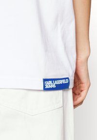 Hvid t-shirt lavet af bomuld, med et blåt mærke i kanten hvor der står "KARL LAGERFELD JEANS" i hvid tekst. Enkel design, lige snit.