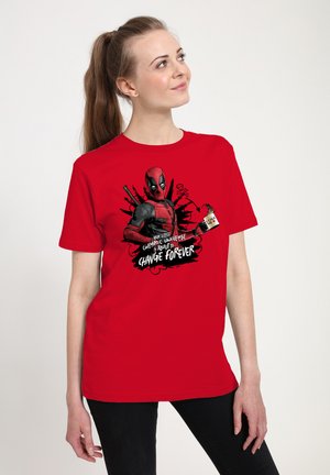 Femme portant un t-shirt Deadpool rouge avec un visuel et un texte sur le changement de l'univers cinématographique, debout sur un fond blanc.