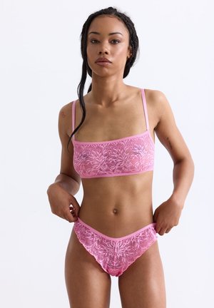 Vrouw in een roze kanten tweedelig lingerieset, die de onderkant aanpast, met gevlochten haar en een neutrale uitstraling tegen een witte achtergrond.