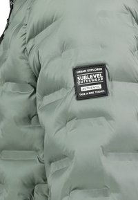 Helle grüne Puffjacke mit schwarzem quadratischem Aufnäher, auf dem steht: "Urban Explorer Sublevel Outerwear Authentisch Riskieren Sie heute!"
