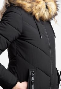 Chaqueta de invierno acolchada negra con cremallera frontal, capucha con borde de piel sintética y detalles de bolsillo con cordón ajustable.