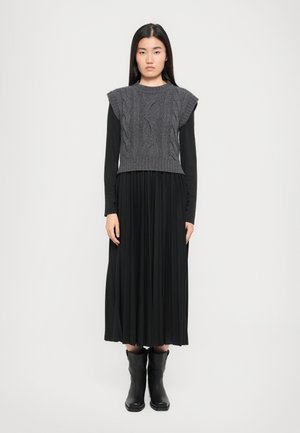 Chaleco de punto gris con patrón de trenzas sobre un vestido negro largo con pliegues, acompañado de botas negras hasta el tobillo. Diseño simple y monocromático.