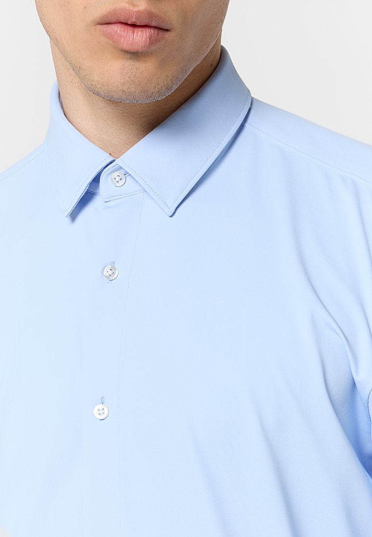 Chemise bleu clair à boutons avec un col structuré, présentant une texture lisse et deux boutons visibles sur la patte de boutonnage avant.