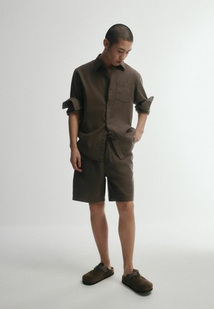 Mann trägt braunes Hemd mit Knöpfen und hochgekrempelten Ärmeln, passende Shorts und braune Slipper, steht vor einem einfarbigen hellen Hintergrund.