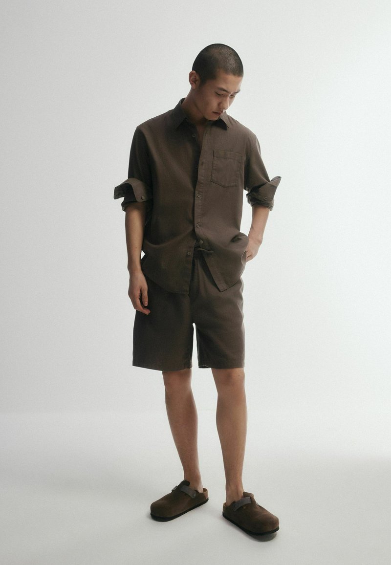 Homme portant une chemise marron à boutons avec manches retroussées, un short assorti et des chaussures marron sans lacets, debout devant un fond clair uni.