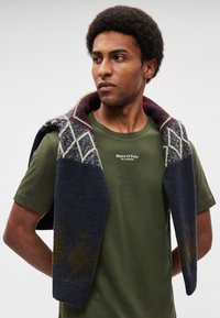 Μερικό ναυτικό πουλόβερ πλεκτό με διαμάντι μοτίβο και μαρουν τόνους, που ακουμπά πάνω σε ένα βαμβακερό T-shirt σε ελιά πράσινο χρώμα. Μαλακή υφή.