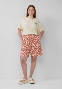 T-shirt crema a maniche corte con una grafica del sole, abbinata a pantaloni corti marrone rossiccio con un motivo astratto in crema chiaro. Sandali platform spessi.