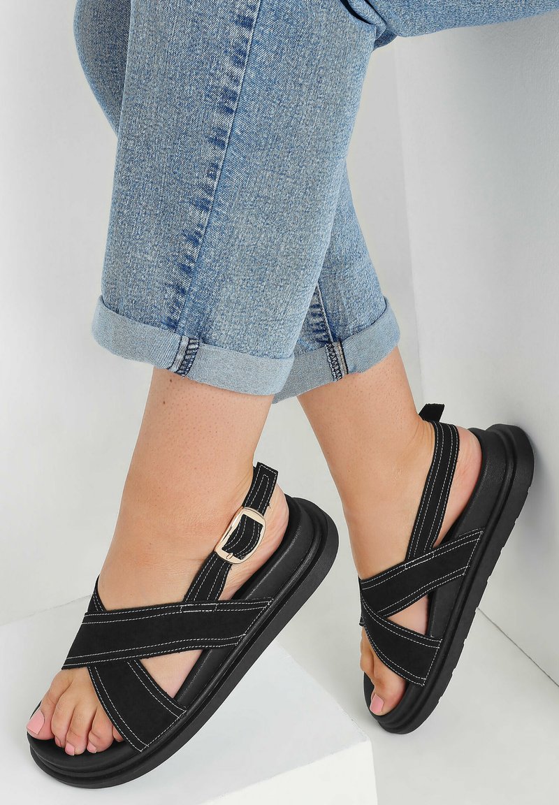 Mustat sandaalit, joissa on ristikkäinen etumuotoilu, varustettuna solkiremmeillä ja valkoisilla ompeleilla. Teksturoitu pohja ja avonainen varvasosa. Yhdistettynä käännettyihin farkkuliiveihin.