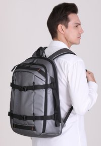 Zaino grigio con spallacci neri, multiple tasche e un accento arancione. Realizzato in materiale resistente, presenta un design elegante e rettangolare.