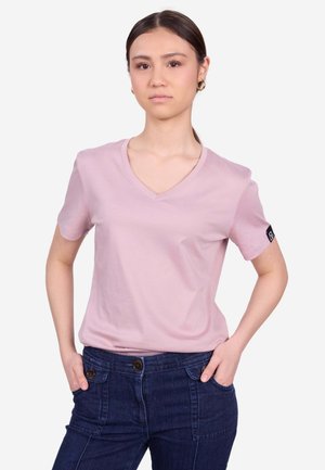 Giovane donna con capelli scuri che indossa orecchini a cerchio dorati, una t-shirt rosa chiaro a scollo a V e pantaloni blu scuro con le mani in tasca.