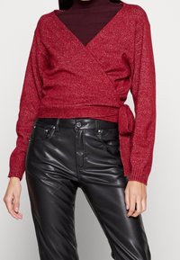 Roter Wickelpullover mit strukturierter Oberfläche, langärmlig und mit einem gebundenen Taillendetail, kombiniert mit schwarzen Kunstlederhosen.