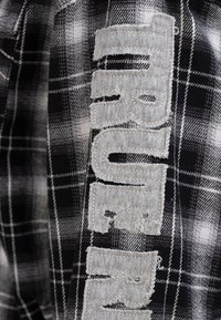 Czarna i biała tkanina w kratę z szarym wyhaftowanym napisem "TRUE". Materiał widoczny z teksturowanym wykończeniem i postrzępionymi krawędziami na literach.