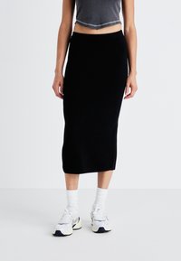 Jupe tricotée noire, longueur genou, au design ajusté, assortie à des baskets blanches et des chaussettes mi-mollet. Le tissu semble doux et extensible.