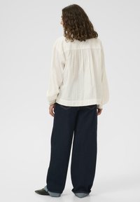 Femeie cu păr creț, îmbrăcată cu o bluză albă cu mâneci lungi și blugi evazați de culoare albastră închisă, stând cu spatele la un fundal simplu.