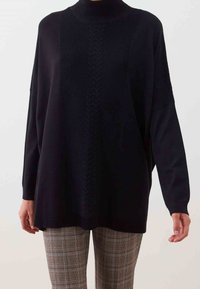 Czarny, oversize'owy sweter z wysokim kołnierzem i prążkowaną fakturą, z centralnym wzorem warkoczowym. W zestawie z kraciastymi spodniami.