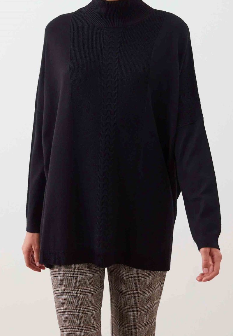 Czarny, oversize'owy sweter z wysokim kołnierzem i prążkowaną fakturą, z centralnym wzorem warkoczowym. W zestawie z kraciastymi spodniami.