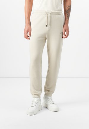 La modelo lleva pantalones de chándal beige con una cinturilla elástica y puños, combinados con zapatillas blancas, todo ello contra un fondo blanco liso.