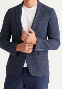 Muž upravující manžetu texturovaného tmavě modrého saka nošeného přes bílé tričko s kulatým výstřihem, tmavě modré kalhoty, stříbrný prsten a tenký řetízkový náhrdelník.