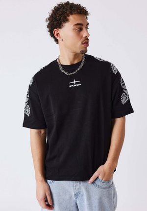 Uomo con capelli ricci che indossa una maglietta nera con disegni floreali bianchi sulle maniche e logo sul petto, jeans azzurri chiari e collana d'argento.