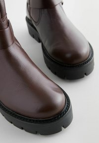 Botas de piel marrón hasta el tobillo con un acabado texturizado, punta redondeada, suela gruesa de goma negra y costura detallada a lo largo del borde.