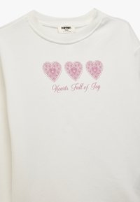 Sudadera blanca de algodón con tres diseños de corazones rosa con detalles de encaje y el texto "Corazones Llenos de Alegría" debajo.