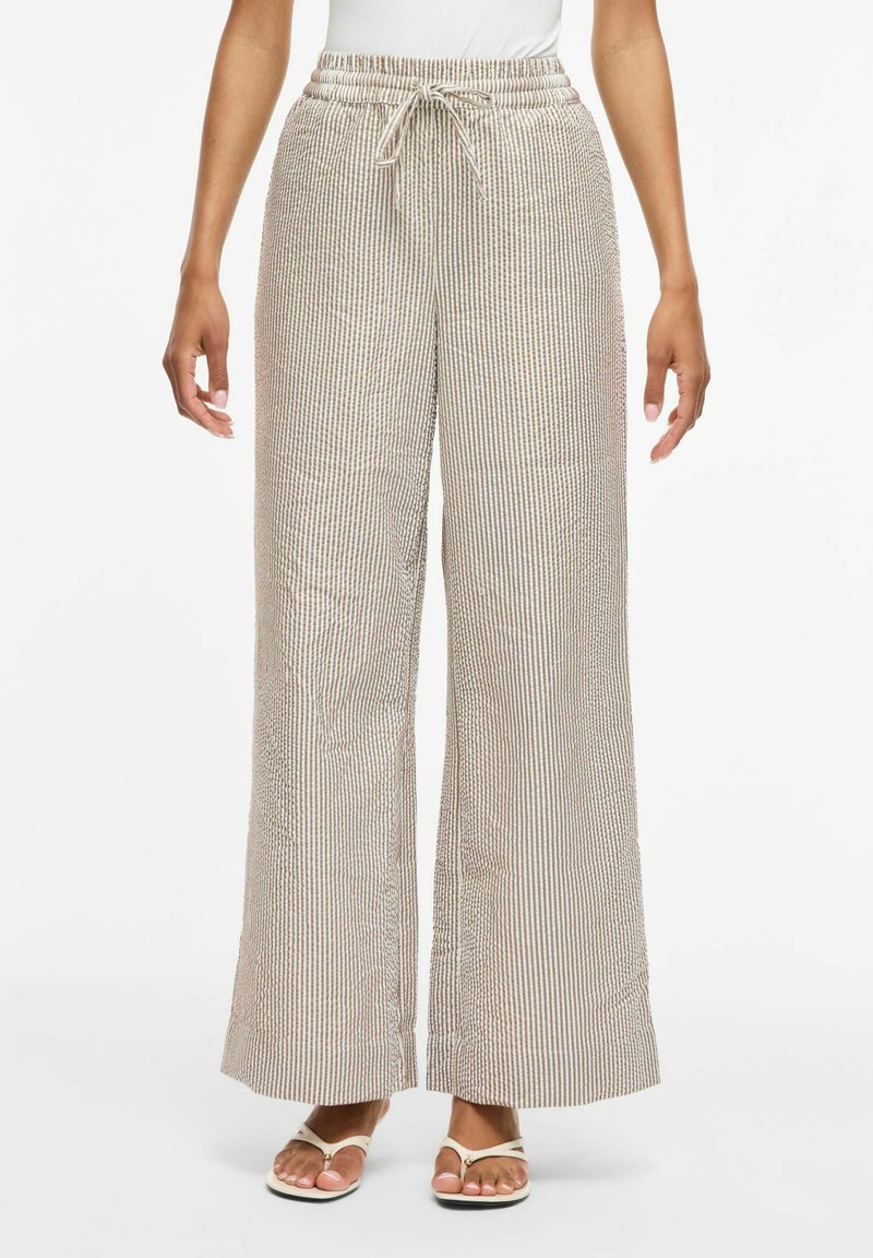 Femme portant un pantalon large beige et blanc à rayures avec une taille élastiquée et des sandales blanches type tongs, debout devant un fond blanc.