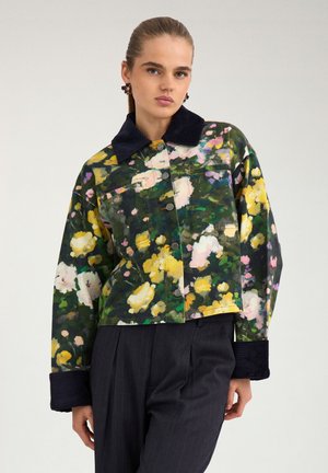 Femme portant une veste fleurie boutonnée avec des fleurs vertes, jaunes et roses, un col et des poignets en velours côtelé noir, et un pantalon plissé foncé.