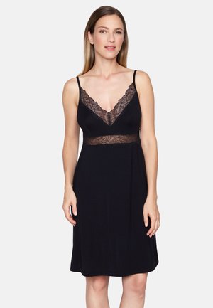 Kobieta ubrana w czarną, bezrękawnikową sukienkę z koronkowym wykończeniem na dekolcie i talii, stojąca na jednolitym jasnym tle.