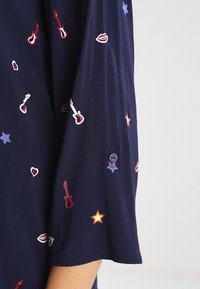 Haut en tissu bleu marine avec des manches courtes mettant en avant une broderie colorée de guitares, cœurs, lèvres et étoiles. Comprend un détail de bouton sur la manche.
