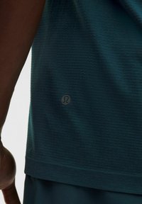 Teal színű sportpóló, texturált anyagból készült, rövid ujjú, és egy kis fényvisszaverő logó található az alján.