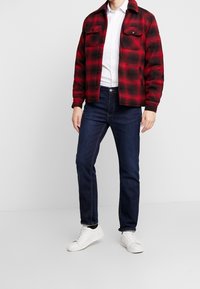 Veste en flanelle à carreaux rouge et noir avec deux poches poitrine, chemise blanche en dessous, jean bleu foncé et baskets blanches. Tenue décontractée.