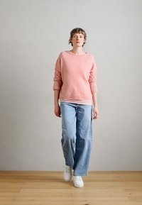 Camisola cor-de-rosa com mangas compridas, usada sobre uma t-shirt branca; combinada com jeans largos azul-claro e ténis brancos, num fundo neutro.