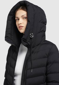 Jachetă puffer neagră cu o glugă mare, dotată cu fermoar și detalii cu șnur. Materialul pare căptușit și neted.