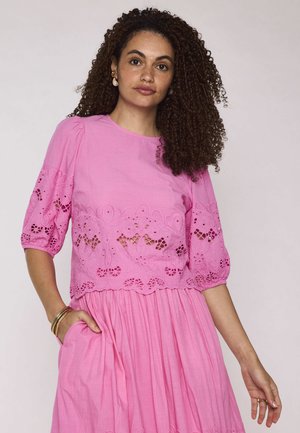 Donna con capelli ricci che indossa un vestito rosa con ritagli ricamati, posa con una mano in tasca e un'espressione neutra su sfondo chiaro.