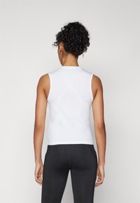 Hvit ermeløs topp med høy hals, laget av et glatt stoff. Baksiden er enkel med en tettsittende design, kombinert med sorte leggings.