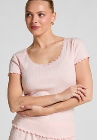 Korte mouwen ribben top in lichtroze met gegolfde randen en een ronde halslijn. De stof heeft een textuurpatroon dat subtiele diepte creëert.