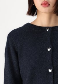 Marineblauwe wollen cardigan met een ronde halslijn, voorzien van vier metalen hartvormige knopen aan de voorkant. Zachte textuur en een ontspannen pasvorm.