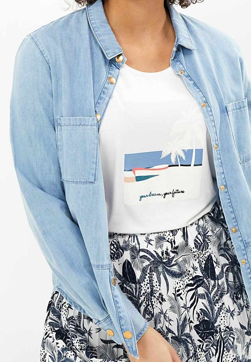 Chemise en denim bleu clair avec des boutons dorés portée sur un t-shirt blanc à motif plage. Jupe fleurie imprimée en noir et blanc.